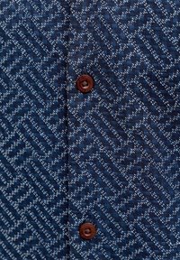 Σκούρο μπλε ύφασμα με περίπλοκη λευκή γεωμετρική ραφή και δύο καφέ κουμπιά ραμμένα κάθετα κατά μήκος μιας κεντρικής ραφής.