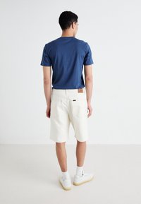 T-shirt en coton marine à manches courtes, associé à un short en denim blanc avec une étiquette en cuir. Le mannequin porte des baskets blanches et des chaussettes.