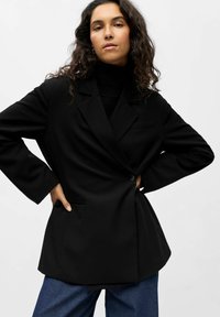 Juodas švarkas su pritaikytu kirpimu, notch tipo atlapais, vieninteliu mygtuku ir šoninėmis kišenėmis. Dėvimas ant juodo megztinio su aukštu kaklu ir derinamas su mėlynais džinsais.