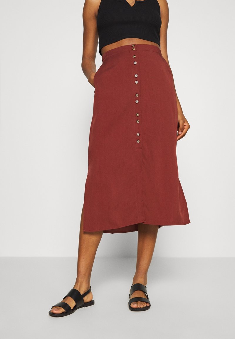 Femme portant une jupe midi boutonnée couleur rouille, un top court noir sans manches et des sandales noires, debout devant un fond uni.