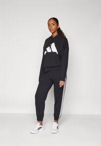 Svart kortermet hettegenser med hvit logo, kombinert med svarte joggebukser med hvite sidestrekker. Modellen har på seg hvite treningssko.