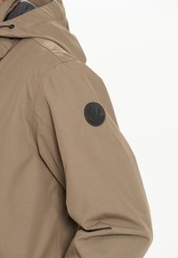 Beige jacka med ett strukturerat, vattenavvisande tyg. Har en svart logotyp patch på vänster axel och en justerbar huva.