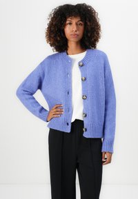 Lyse lilla strikket cardigan med rund hals, seks knappelukkinger og ribbede mansjetter, kombinert med en hvit skjorte og mørke bukser.