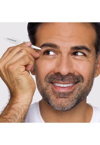Uomo con barba corta che sorride mentre si sistema il sopracciglio con pinzette argento del marchio Tweezerman su sfondo semplice.