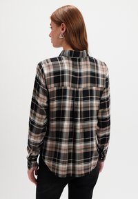 Kobieta z długimi czerwonymi włosami, ubrana w beżową, czarną i szaro-koszulową koszulę, srebrne trójkątne kolczyki i czarne spodnie, odwrócona plecami na białym tle.