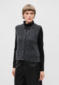 Tumehallid kootud vest tekstuuriga viimistlusega, ümarate kaeluste, viie musta nööbi ja varrukateta disainiga musta särke peal.