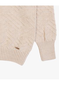Maglione lavorato a maglia beige con motivo a spina di pesce, polsini a coste e una piccola etichetta decorativa sul lato. Texture morbida, peso medio.