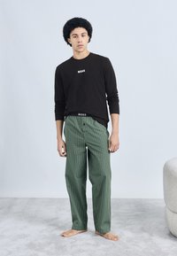 Cămașă neagră cu mâneci lungi cu logo-ul "BOSS", asortată cu pantaloni de pijama verde cu carouri. Material moale și croială lejeră, picioarele goale vizibile.