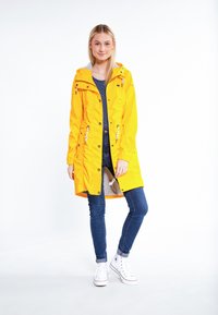 Chaqueta impermeable amarilla con capucha, cintura con cordón, bolsillos con cremallera, combinada con una blusa gris a rayas, vaqueros azules y zapatillas blancas.