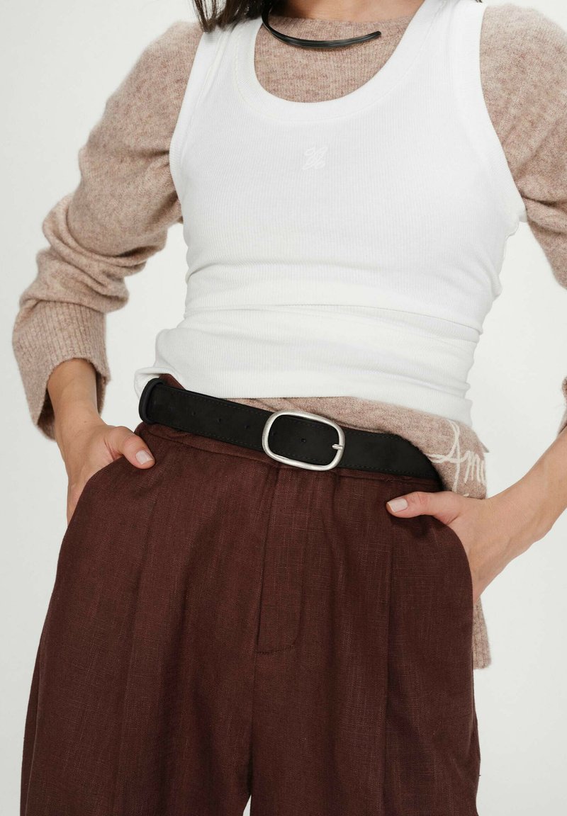 Personne portant un pull à manches longues marron superposé sous un débardeur blanc, pantalon marron foncé avec ceinture noire, mains dans les poches.