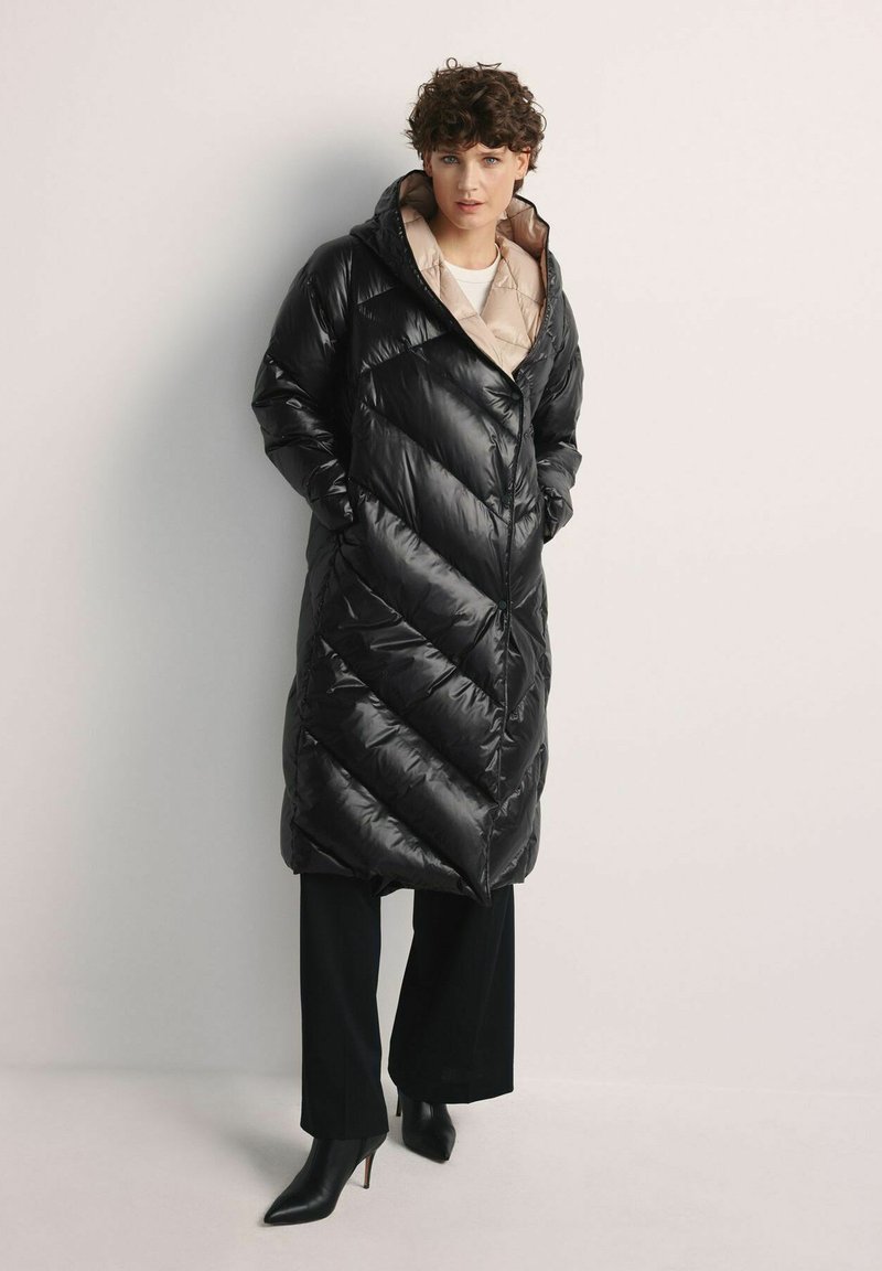 Czarna kurtka puchowa z wzorem w zygzak, kapturem i beżową podszewką, zestawiona z szerokimi spodniami i czarnymi szpilkami.