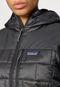 Črno podloženo jakno s kapuco, z sprednjim zadrgo in logotipom Patagonia. Tekstura je gladka z prešito površino.