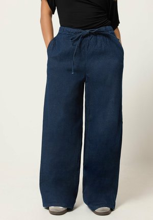 Mujer con pantalones vaqueros de pierna ancha de tono azul oscuro, con cintura elástica y cordón, combinados con zapatos grises y camiseta negra. Manos en los bolsillos.