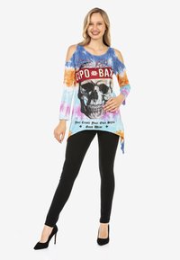 Langärmliges Tie-Dye-Oberteil mit ausgeschnittenen Schultern, das eine Schädelgrafik und Text zeigt. Kombiniert mit schwarzen engen Hosen und schwarzen High Heels.