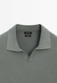 Polo en maille texturée gris clair avec col ouvert et étiquette noire cousue à l’intérieur de l’encolure.