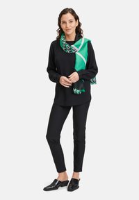 Czarna bluzka z długim rękawem, dopasowane czarne spodnie i zielony szalik w geometryczne wzory. Modelka stoi z lekko zginionymi ramionami.