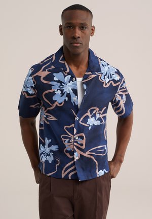 Muž nosi tmavomodrú košeľu s krátkym rukávom na gombíky s veľkými svetlomodrými a ružovými kvetinovými vzormi, v kombinácii s hnedými nohavicami.