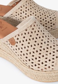 Zapatos slip-on de ante beige con patrón geométrico recortado, suela de cuña envuelta en yute y etiqueta de cuero. Textura suave en toda la superficie.