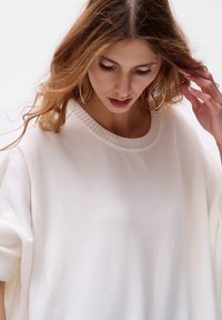 Overmaat wit gebreid sweater met geribbelde halslijn en korte mouwen. Zachte textuur, minimalistisch ontwerp en iets losse pasvorm.