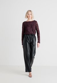 Burgundska teksturirana majica s dugim rukavima s složenim uzorkom, u paru s crnim, slobodno krojenim hlačama, stvara ležeran, ali profinjen izgled.