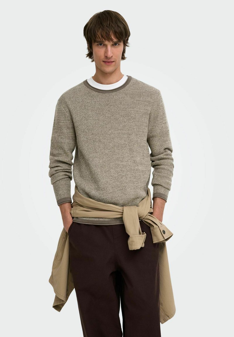 Jeune homme aux cheveux bruns portant un pull beige par-dessus une chemise blanche et un pantalon foncé, avec une veste beige nouée autour de la taille.