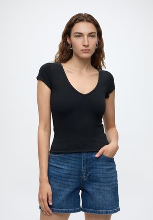 Jeune femme aux longs cheveux ondulés portant un haut noir à manches courtes en V et un short en jean bleu, debout avec une main dans la poche.