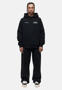 Hoodie nero oversize con tasca a marsupio, caratterizzato da stampe di testo bianche. Abbinato a pantaloni felpati neri larghi e sneakers nere.