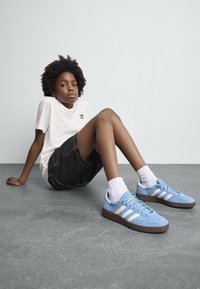 Modri gamisnki športni copati z belimi črtami in rjavimi gumijastimi podplati. Model nosi belo majico in črne kratke hlače, seden na sivi površini.