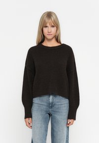 Mørkebrun strikkegenser med løs passform, vide ermer og rund hals, sammen med lyseblå denimjeans.
