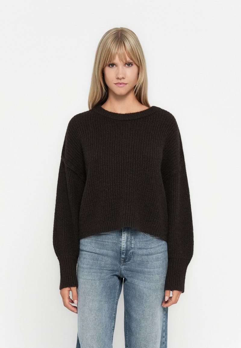 Mørkebrun strikkegenser med løs passform, vide ermer og rund hals, sammen med lyseblå denimjeans.