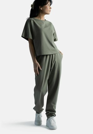 Nainen seisoo, päällään väljät oliivinvihreät paita ja housut sekä valkoiset lenkkarit, katsoo sivulle suorassa vaaleassa taustassa.