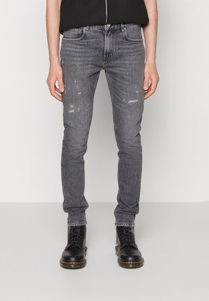 Mann trägt schlanke graue Jeans mit Used-Look und schwarzen Schnürstiefeln, steht vor einem schlichten hellen Hintergrund.