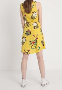 Gelbes ärmelloses Kleid mit floralem Muster, ausgestattet mit einem elastischen Bund und einem ausgestellten Rock. Getragen mit weißen High-Top-Sneakern.