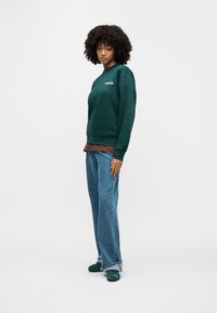 Γυναίκα με σγουρά μαλλιά που φοράει σκούρο πράσινο φούτερ, καφέ μπλουζάκι, φαρδιά μπλε τζιν και πράσινα παπούτσια Puma, στέκεται μπροστά σε λευκό φόντο.