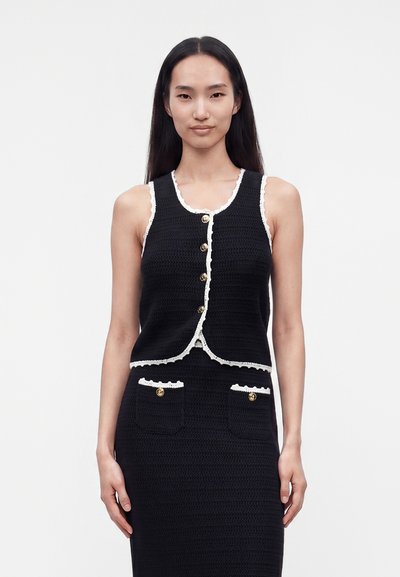 Femme portant un gilet noir sans manches boutonné avec bordure blanche et une jupe noire assortie avec deux poches avant et des boutons dorés.