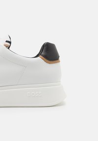 Weißer Sneaker mit strukturiertem Sohlen und dem eingeprägten "BOSS"-Logo an der Seite, versehen mit schwarzen und beige Lederakzenten an der Ferse.