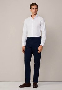 Hvid langærmet skjorte med knapper kombineret med mørkeblå bukser og brune lædersko. Pasformen er skræddersyet, hvilket skaber en pæn silhuet.