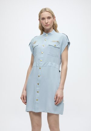 Frau mit hellem Haar, die ein hellblaues Kleid mit Knopfleiste, goldenen Knöpfen und zwei Brusttaschen trägt, vor einem schlichten weißen Hintergrund steht.