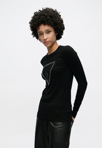 Μαύρο πουλόβερ με μακριά μανίκια που διαθέτει τριγωνικό λογότυπο με μεταλλική περίμετρο, κατασκευασμένο από ομαλή, ελαφριά υφή.