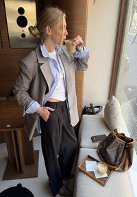 Femme en blazer superposé et pantalon boit un café près de la fenêtre dans un café avec un sac, un téléphone et un plateau avec un latte sur un banc rembourré.