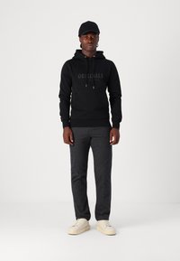 Sudadera negra con el logo "ORIGINALS", pantalones grises texturizados y zapatillas blancas. El modelo lleva una gorra negra y está de pie contra un fondo liso.