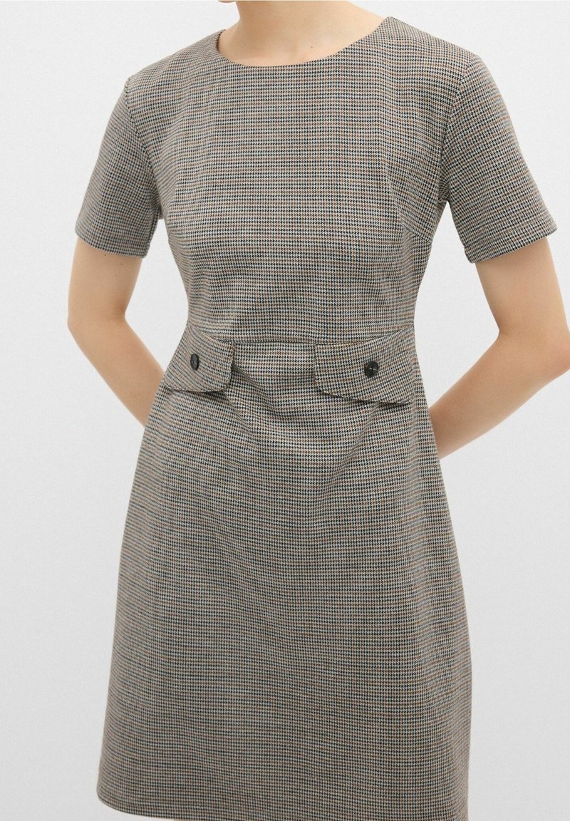 Femme portant une robe courte à manches courtes, longueur genou, avec un petit motif à carreaux et deux poches avant à rabat avec des boutons noirs.