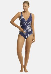 Model w granatowym kostiumie kąpielowym w kwiaty, uśmiechająca się i poprawiająca dopasowanie, na jednolitym białym tle. Na boso, prezentująca zrelaksowaną postawę.