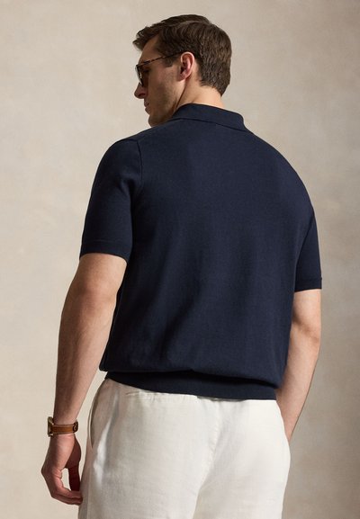 Άνδρας που φοράει σκούρο μπλε κοντομάνικο πόλο και άσπρο παντελόνι, από πίσω, με γυαλιά ηλίου και καφέ δερμάτινο ρολόι.