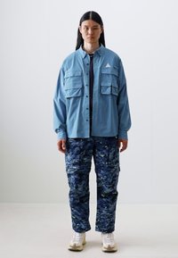 Lys blå skjorte med knapper og flere brystlommer, paret med marineblå cargo-bukser med camo-mønster. Skoene er hvite med beige detaljer.