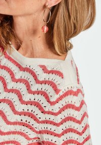 Strickpullover mit weißem Hintergrund und korallenfarbenem Zickzackmuster. Verfügt über einen weiten Halsausschnitt und eine offene Webstruktur. Große Creole mit rosa Perle.