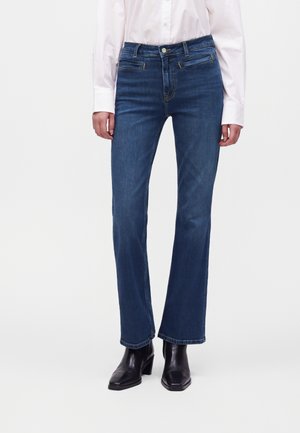 Femme portant un jean droit bleu foncé, une chemise blanche à manches longues rentrée, et des bottines noires, debout devant un fond uni.