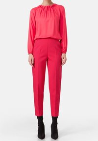 Vrouw in een felroze blouse met lange mouwen, ingestopt in bijpassende roze, nauwsluitende hoge taille broek, gecombineerd met zwarte enkellaarsjes.