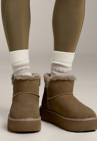 Braune Wildleder-Stiefeletten mit Fellbesatz, getragen mit weißen gemusterten Wollsocken und olivgrünen Leggings vor einem neutralen Hintergrund.