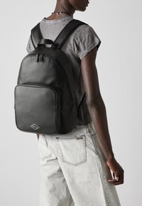 Zaino in pelle nera con una texture liscia, dotato di una tasca frontale con zip e di un logo a diamante. Indossato sulla schiena, abbinato a un abbigliamento grigio.